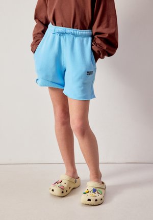 Inimene, kes kannab helesiniseid fliisist lühkseelikke, millel on kiri "American Vintage", pruunid pika varrukaga pluusi ja beežid sandaalid dekoratiivsete ehetes.