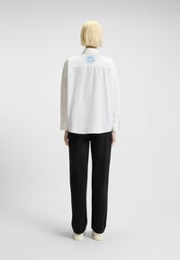 Witte blouse met een blauwe geborduurde zon aan de achterkant, gecombineerd met zwarte broeken. Gladde stof, losse pasvorm, lange mouwen, knoopmanchetten.
