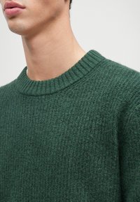 Mørkegrøn striktrøje med struktureret overflade og ribbet rund halsudskæring. Stofferne ser varme og hyggelige ud.