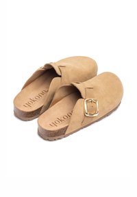 Tan suede instappers met een brede band en een gouden gesp, een getextureerde kurken zool en een gladleren binnenzool.