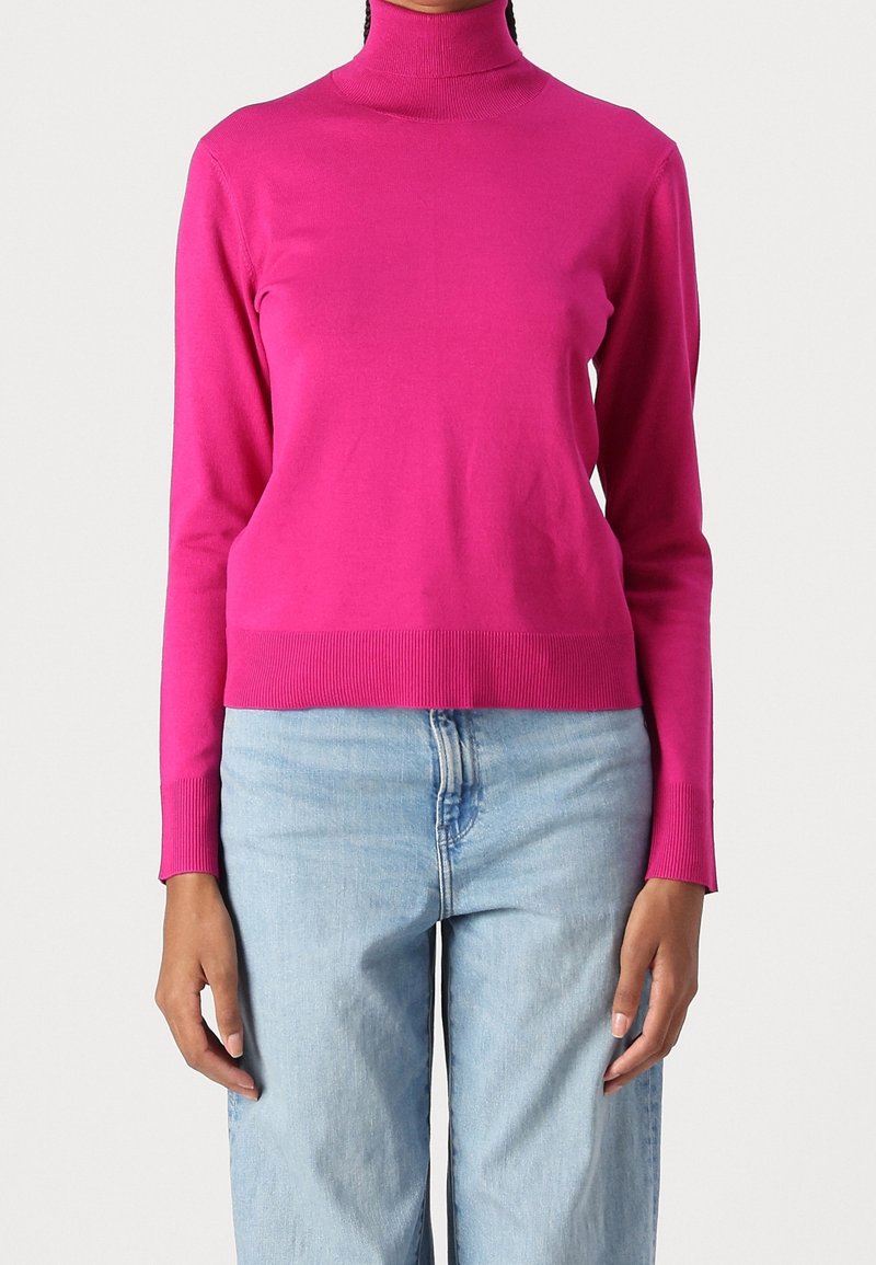 Femme portant un pull à col roulé rose vif et un jean taille haute bleu clair, debout devant un fond blanc uni.