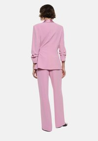 Completo su misura rosa con blazer aderente e pantaloni a gamba larga. Presenta spalle strutturate, una texture liscia e una leggera lucentezza.