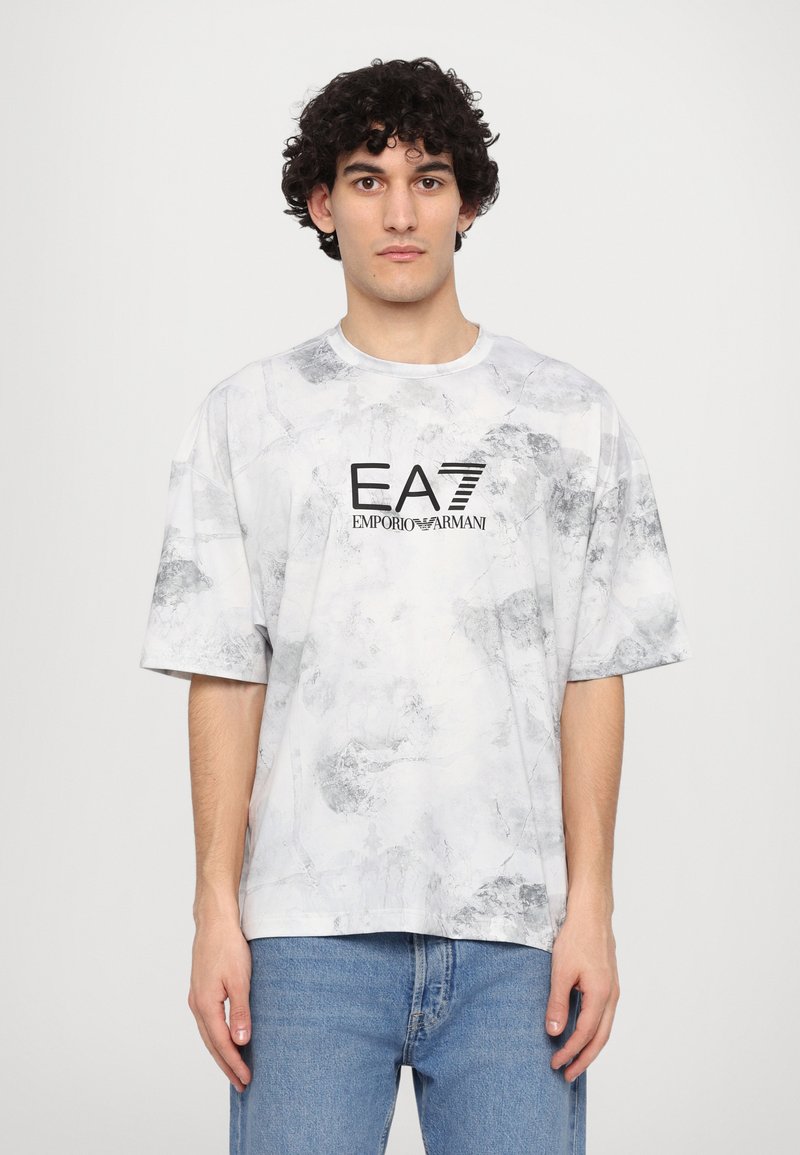 Camiseta blanca con patrón de mármol, de cuello redondo y manga corta. Presenta un logo negro de EA7 en la parte delantera. Combinada con jeans azules.