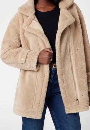 Personne portant un manteau beige duveteux par-dessus un haut noir et un jean bleu, tenant le devant du manteau près de la fermeture éclair.
