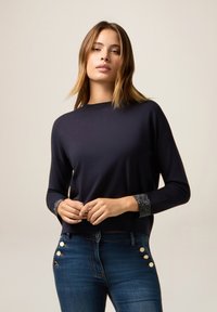 Jonge vrouw die een donkerblauwe trui met versierde manchetten en hoge blauwe spijkerbroeken met decoratieve knopen draagt, terwijl ze tegen een effenberg staat.