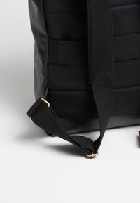 Schwarze Tasche mit glatter, gummierter Textur, verstellbarem Nylontragegurt und Akzenten in Goldfarbe. Seitenansicht zeigt vertikale Nähte.