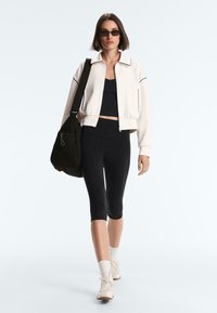 Nainen, jolla on aurinkolasit, valkoinen vetoketjullinen takki, musta lyhyt paita, mustat capri-leggingsit, valkoiset lenkkarit ja suuri musta olkalaukku.