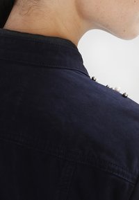 Marinblå skjortkrage med blanka dekorativa pärlor i svart och vitt vid axeln. Mjuk textur med en subtil lyster.