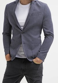 Mörkblå texturerad kavaj med en figursydd design, enfaldig knäppning, slagkrage och två framfickor, bärs över en vit skjorta.