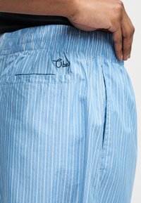 Χέρι ακουμπισμένο στη μέση, φοράει ανοιχτό μπλε ριγέ σορτσάκια με ελαστική μέση και κεντημένο λογότυπο "Obey" πάνω από την πίσω τσέπη.