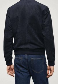 Bomber in tessuto morbido blu navy con polsini e orlo a costine, dal design con finitura liscia. Indossato sopra jeans in denim scuro.