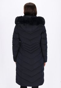 Marineblauwe pufferjas met chevronstiksels, een nepbontkraag, lange mouwen en een knielange pasvorm. Glad oppervlak, warme stof.