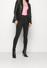 Svart läderblazer över en rosa t-shirt, med svarta skinny jeans och svarta stiletto ankelskor, visad i en stående pose.