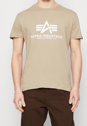 Beżowa koszulka Alpha Industries z białym logo i napisem, noszona przez osobę stojącą z opuszczonymi ramionami, w połączeniu z ciemnobrązowymi spodniami.