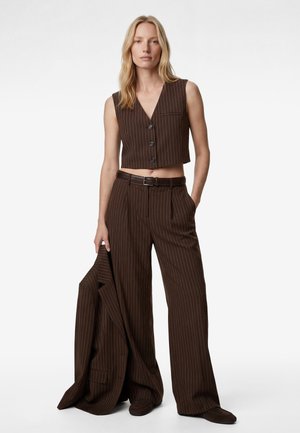 Mujer con chaleco sin mangas corto de raya diplomática marrón y pantalones de pierna ancha sostiene una chaqueta a juego, posando contra un fondo blanco.