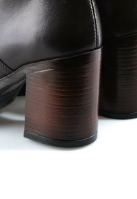 Gros plan de bottes en cuir noir avec des talons blocs en bois épais, empilés et marron foncé sur une surface blanche.
