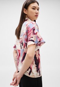 Mladá žena s dlouhými tmavými vlasy, která nosí krátký rukáv halenky s červenými, modrými a béžovými abstraktními vzory, stojící s rukama za zády.