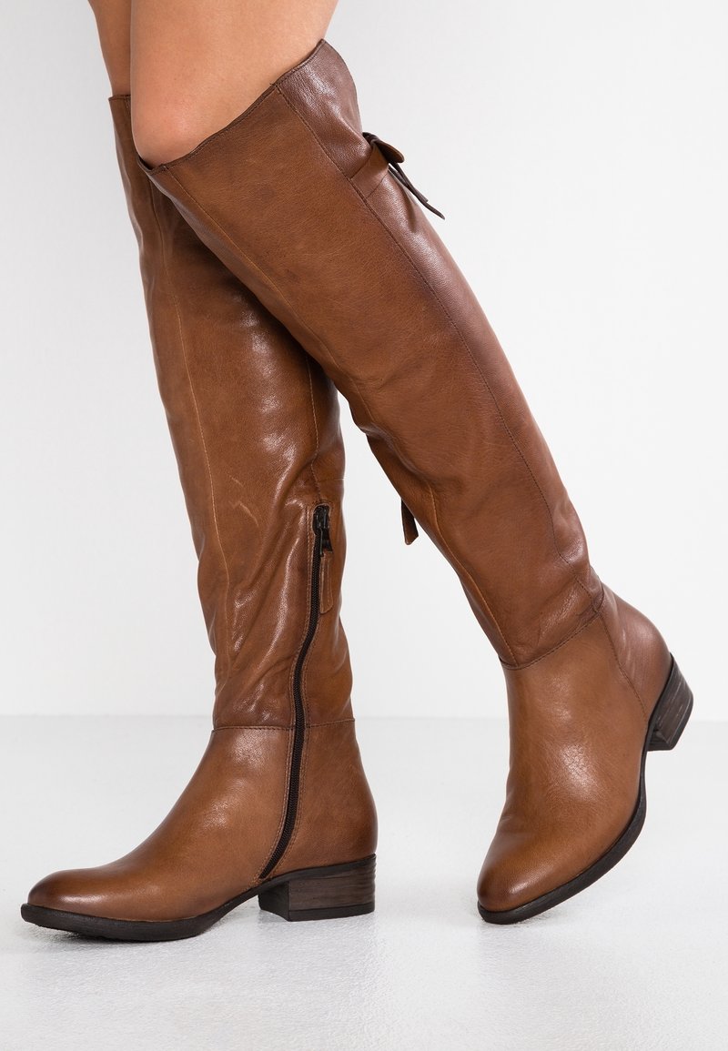 Femme portant des bottes en cuir marron montant jusqu'aux genoux avec une fermeture éclair latérale et un talon bas en bois, debout devant un fond blanc uni.
