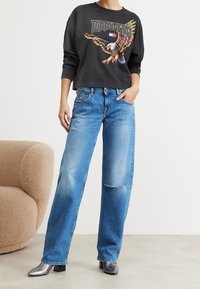 Gesneden zwarte sweatshirt met kleurrijke adelaarafbeelding; hoge taille, lichtblauwe versleten rechte jeans; glanzende zilveren enkellaarsjes.