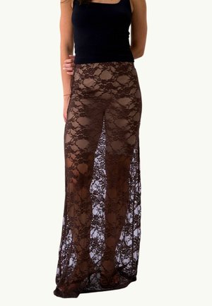 Kvinde iført en lang, gennemsigtig brun blonde nederdel over nude shorts og en sort ærmeløs top, stående mod en ensfarvet baggrund.