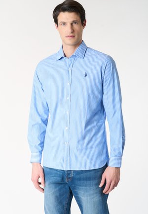 Uomo che indossa una camicia azzurro chiaro a righe verticali con un piccolo logo ricamato sul petto e jeans blu su sfondo bianco.