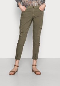 Olive green cargo pants with zipper hems, multiple pockets, and a fitted design. Paired with brown flat sandals.