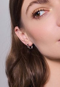 Srebrne kolczyki w kształcie obręczy z twistowanym wzorem, z gładką powierzchnią i błyszczącym wykończeniem. Noszone na uchu modelki, podkreślające ich kształt.