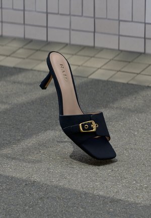 Mule à talons hauts noire avec détail boucle dorée, de la marque "RAID", posée sur une surface asphaltée texturée près d'un pavement carrelé.