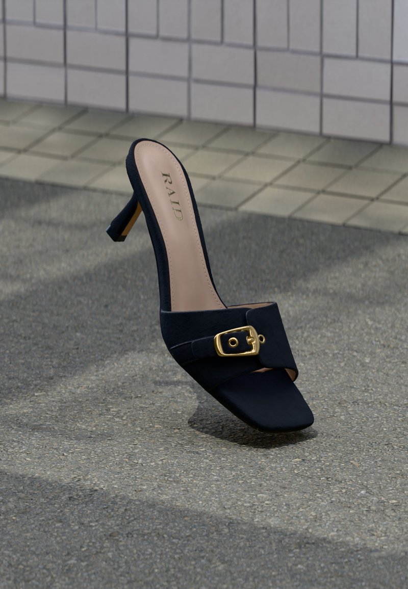 Mule à talons hauts noire avec détail boucle dorée, de la marque "RAID", posée sur une surface asphaltée texturée près d'un pavement carrelé.