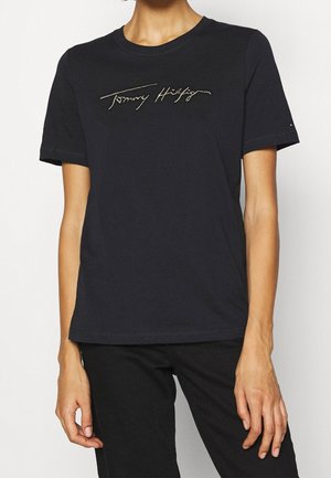 Osoba ubrana w czarną koszulkę z krótkim rękawem z napisem "Tommy Hilfiger" złotą czcionką na piersi, zestawioną z czarnymi spodniami.