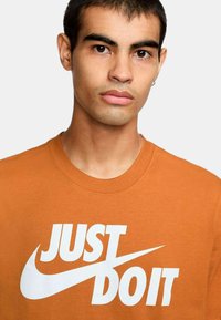 T-shirt in cotone arancione con una grafica bianca audace "JUST DO IT" sul petto, scollo rotondo e maniche corte.