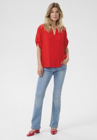 Canottiera rossa a maniche corte con scollatura a V e risvolti, abbinata a jeans azzurri a gamba dritta e scarpe rosse. Tessuto morbido.