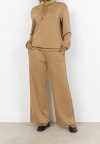 Beżowy dres z bawełny, składający się z bluzy z wysokim kołnierzem i sznurkiem oraz spodni o szerokich nogawkach z kieszeniami. Gładka tekstura i luźny krój.