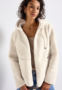 Femme aux cheveux bouclés portant des boucles d'oreilles en or, une veste à capuche beige sur un haut beige, et un jean bleu, posant avec les mains près de la taille.
