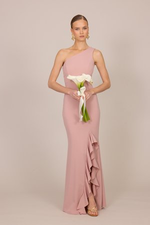 Femme en robe rose une épaule avec un détail de volants tient un bouquet de callas blancs attaché avec un ruban blanc, debout devant un fond uni.