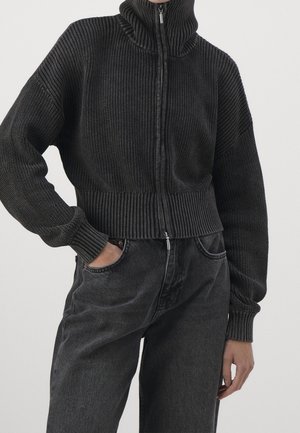 Donkere geribbelde gebreide zip-up trui met een hoge kraag en brede mouwen, gecombineerd met donkere denim jeans. Accenthardware zichtbaar op de rits.