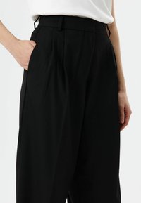 Schwarze, plissierte Hose mit hohem Bund und Seitentaschen; der Stoff wirkt glatt mit leichtem Glanz; schlanke Passform.