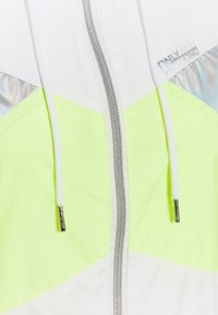 Chaqueta ligera con cremallera en blanco y amarillo neón, con tejido texturizado, detalles reflectantes y capucha con cordón. Detalles en hardware plateado.