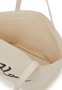 Beige Canvas-Tragetasche mit schwarzem Schriftzug. Geräumiges Innenfach ohne zusätzliche Fächer. Textilhaken zum Tragen.