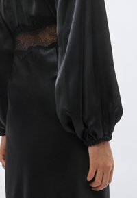 Čierne saténové šaty s dlhými nafúknutými rukávmi. Sú vybavené priesvitným čipkovaným panelom v páse a nariasenými manžetami. Celkovo hladká textúra.