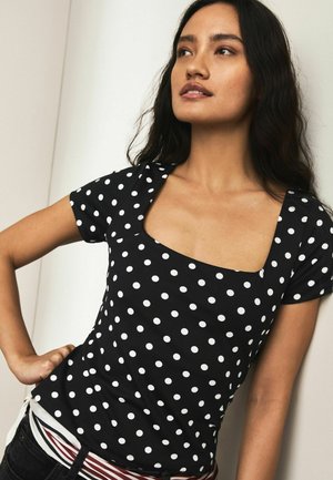 Noor naine, kellel on pikad tumedad juuksed, kannab musta lühikese varrukaga pluusi valgete täppidega, seistes ühe käega puusal.