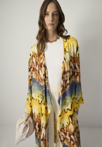 Kimono-tyylinen viitta eläväisessä keltaisessa ja sinisessä abstraktissa kuviossa, kevyt kangas, jossa on väljät hihat, yhdistetty valkoiseen puseroon ja kreemivärisiin housuihin.