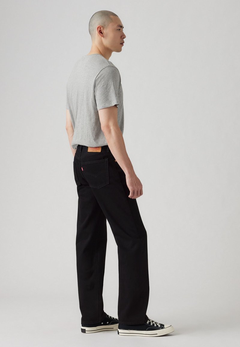 Svarta raka jeans med en synlig bakfickelogga, kombinerat med en ljusgrå kortärmad T-shirt och svarta sneakers.