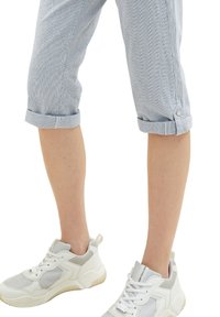 Persona in pantaloni azzurri arrotolati e sneakers bianche in piedi davanti a uno sfondo bianco.