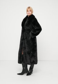 Svart kåpe i kunstpels med sjalskrage, sidelommer og frontåpning. Har en lang, myk tekstur og en slank silhuett.