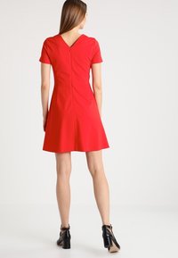 Robe rouge à manches courtes avec un dos en V, jupette évasée et fermeture éclair dissimulée. Tissu lisse sans motifs ni accents visibles.