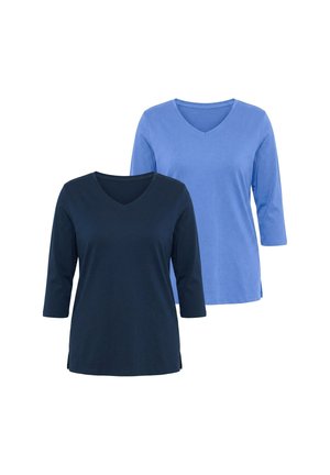 Giovane donna con capelli mossi fino alle spalle, che indossa grandi orecchini a cerchio bianchi, una maglia blu scuro con scollo a V e jeans blu scuro.