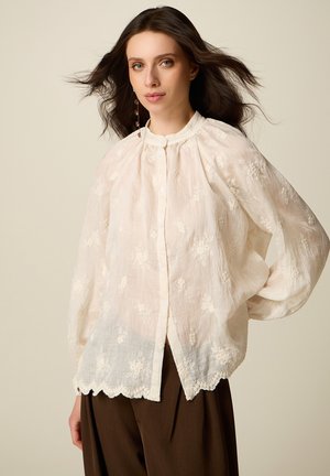 Kvinde med langt mørkt hår iført en hvid broderet bluse med bølget kant og brune bukser, står foran en beige baggrund.