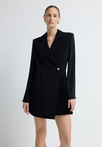 Czarna sukienka o sylwetce dopasowanego żakietu, z długimi rękawami, pojedynczym złotym guzikiem i plisowanym dołem po prawej stronie. Gładki tekstur tkaniny.