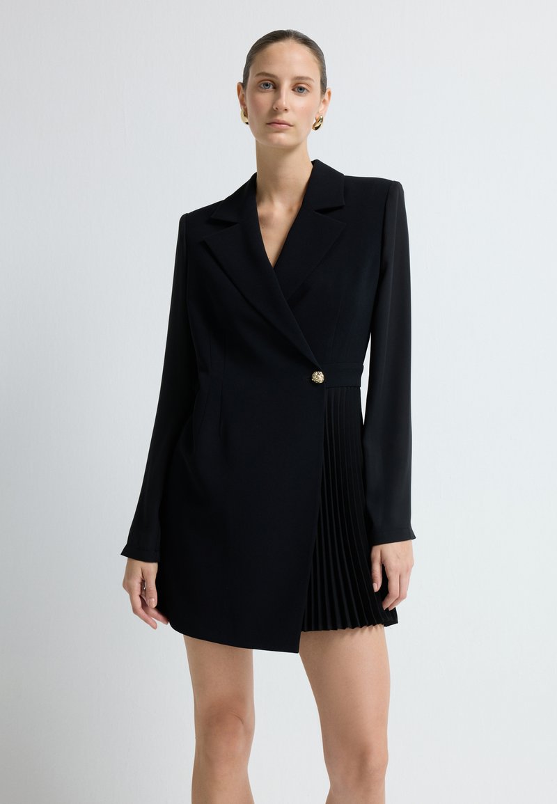 Czarna sukienka o sylwetce dopasowanego żakietu, z długimi rękawami, pojedynczym złotym guzikiem i plisowanym dołem po prawej stronie. Gładki tekstur tkaniny.